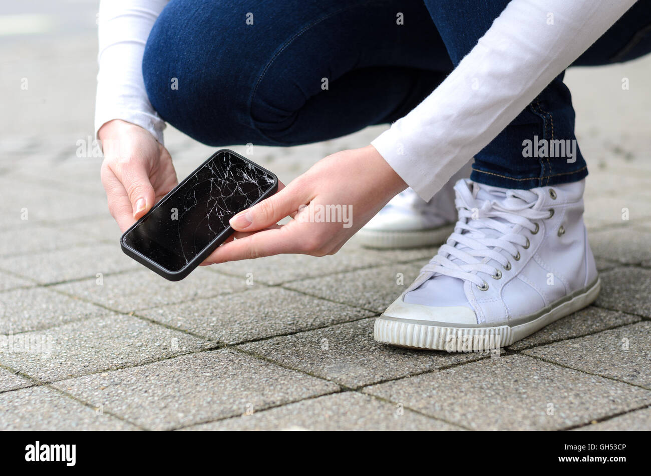 Vista ravvicinata sulla persona in ginocchio in jeans e scarpe di prelevare telefono rotto sul lastricato in pietra marciapiede esterno Foto Stock