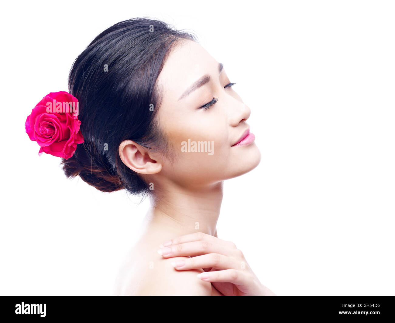 Studio ritratto di una giovane e bella donna asiatica a occhi chiusi, in vista laterale una rosa rossa nei capelli, isolati su sfondo bianco. Foto Stock