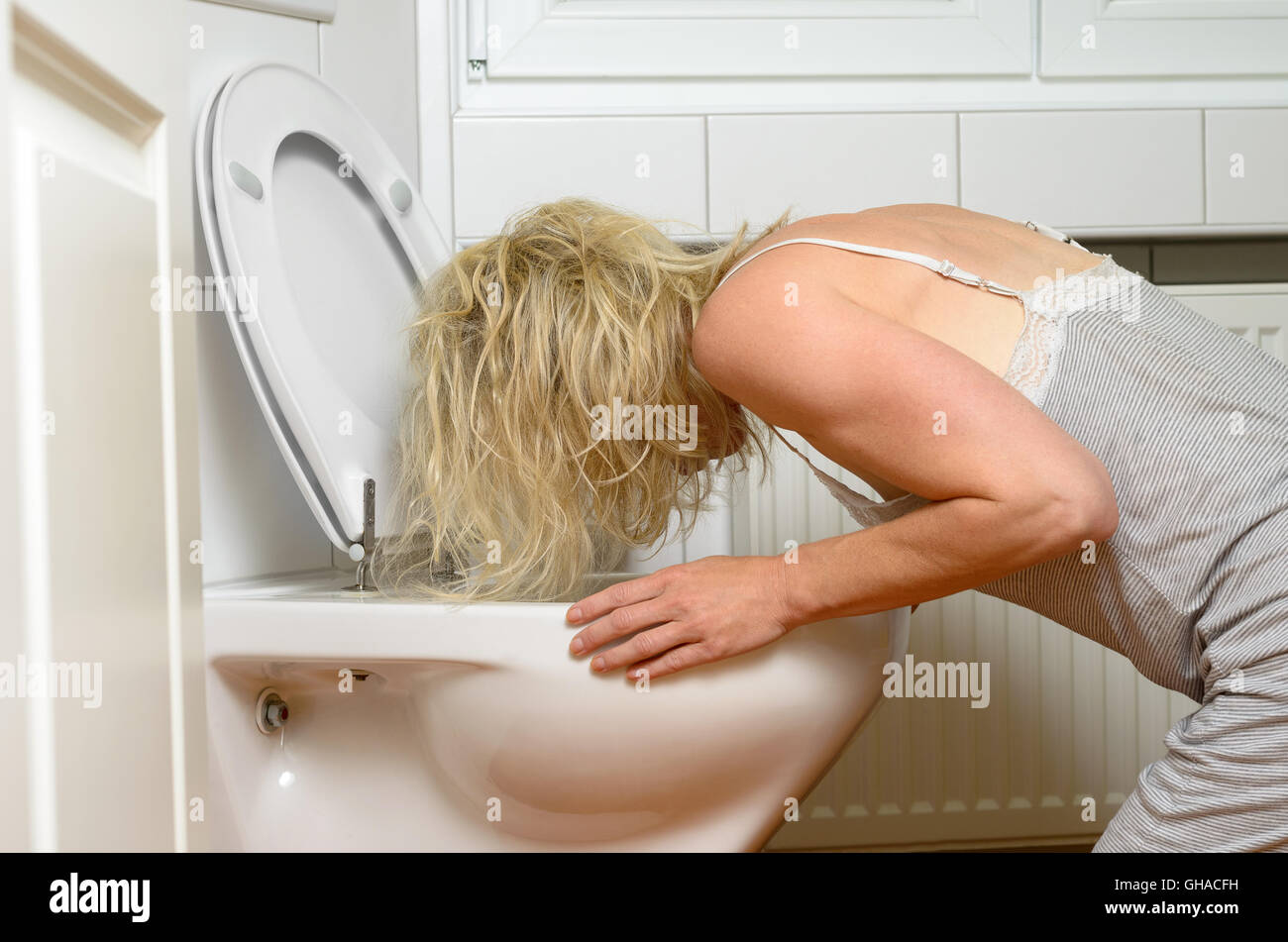 Donna bionda in un abito grigio in ginocchio il vomito in un gabinetto concettuale di una malattia o di un'ubriachezza dopo una festa Foto Stock
