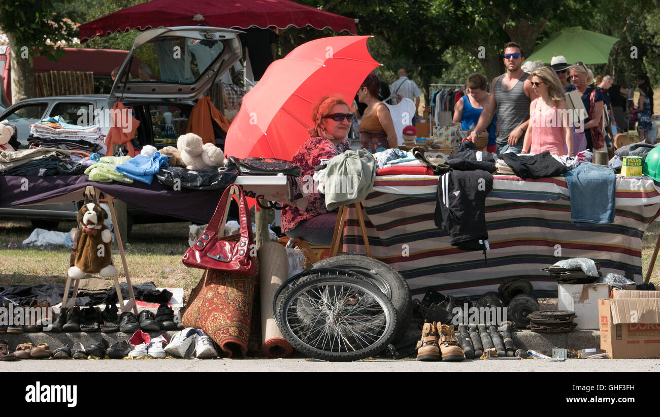 Un venditore al bagagliaio della vettura vendita vide-grenier ripari sotto un ombrello rosso o sun ombra su una torrida giornata di caldo in estate vicino a C Foto Stock