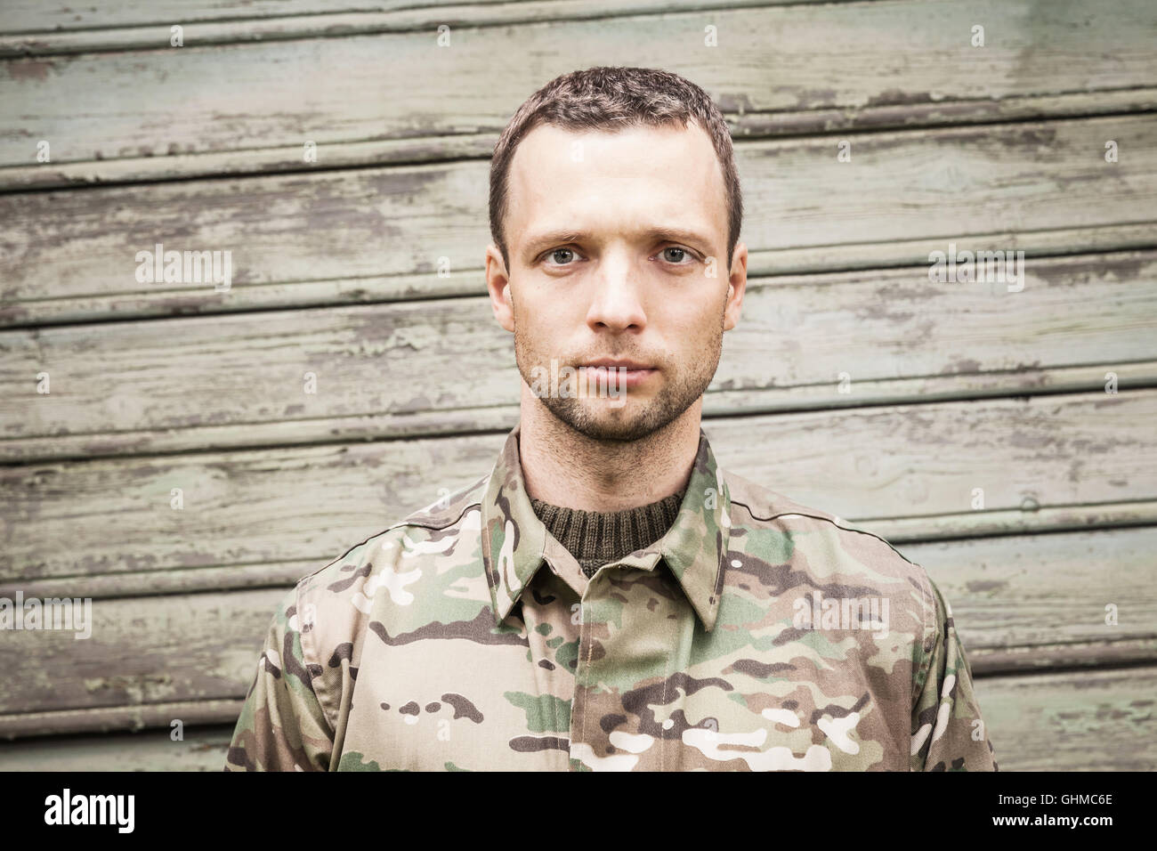 Giovani caucasici grave uomo militare in camuffamento uniforme. Primo piano verticale frontale su verde parete in legno Foto Stock