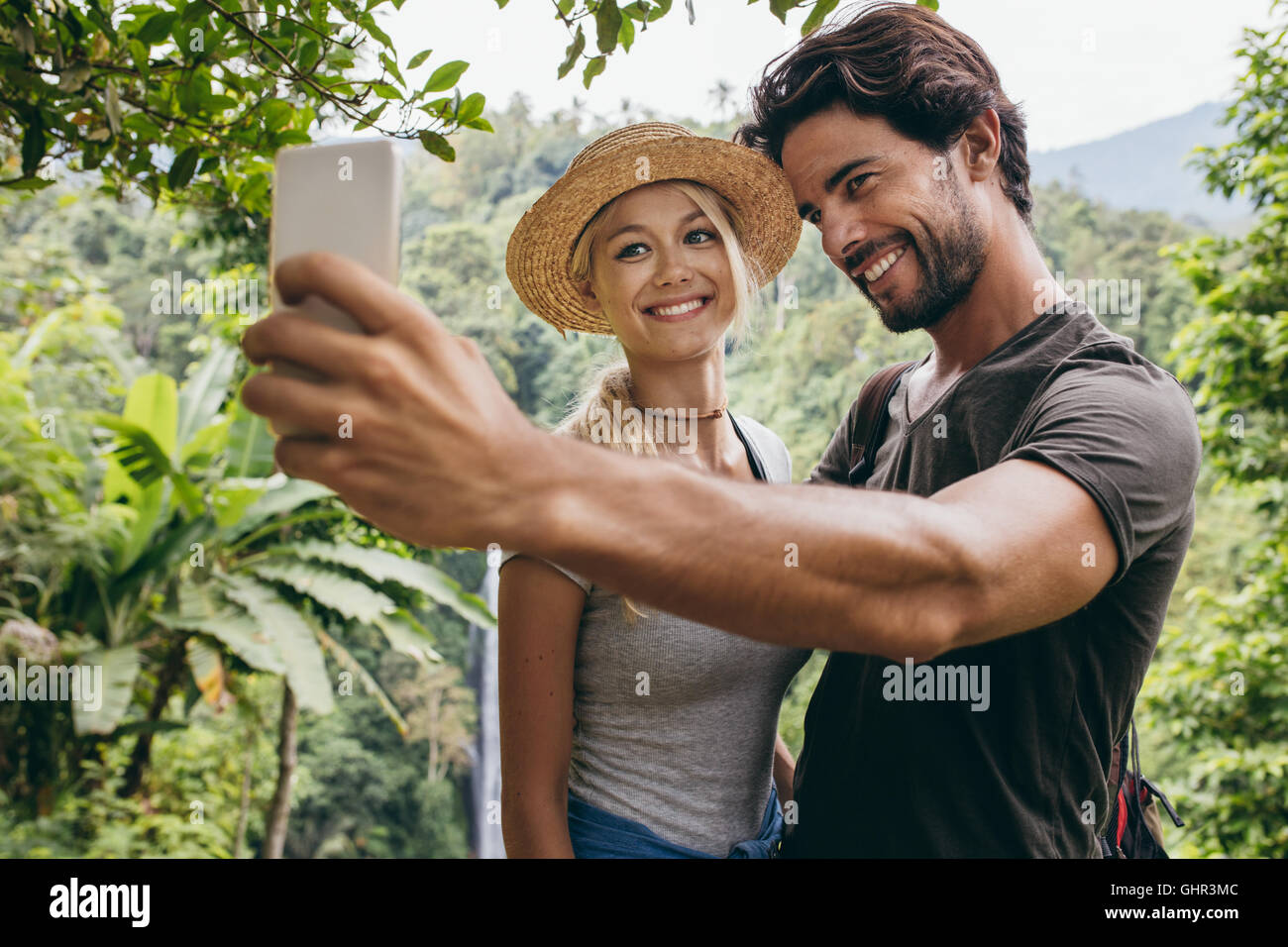 Sorridente coppia giovane tenendo selfie con cascata in background. L uomo e la donna nella foresta tenendo autoritratto con loro cellpho Foto Stock