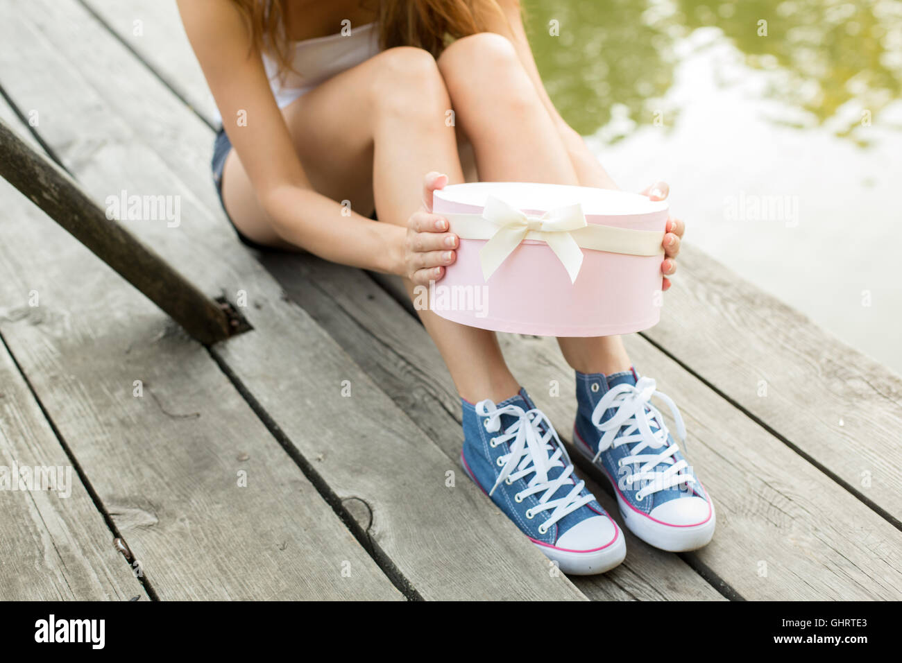 Giovane donna con belle gambe sportivo in jeans sneakers seduto su una apertura di legno di rosa confezione regalo Foto Stock