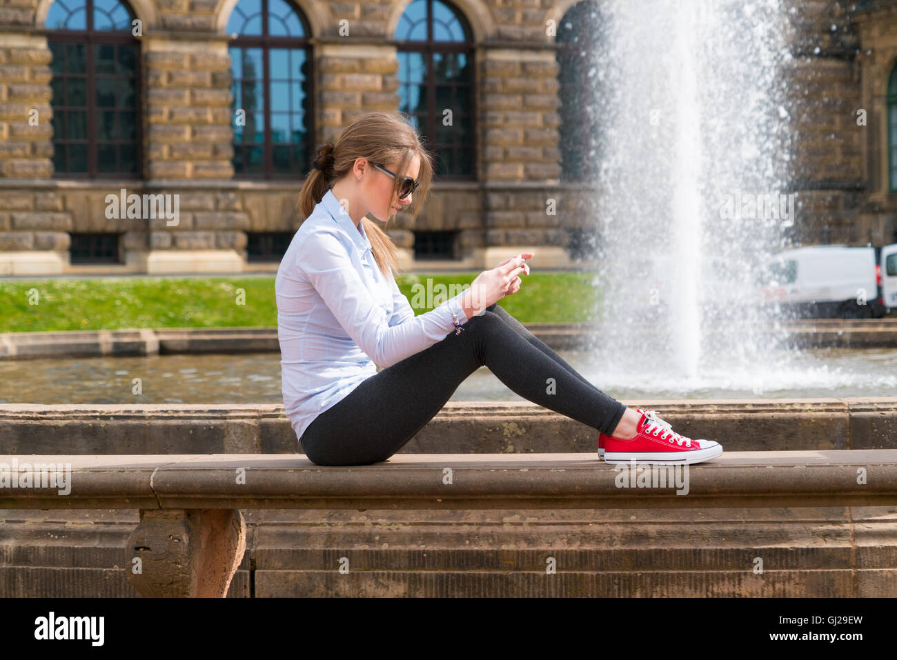 Una giovane donna è seduta da una fontana nella città e sta usando un telefono intelligente Foto Stock