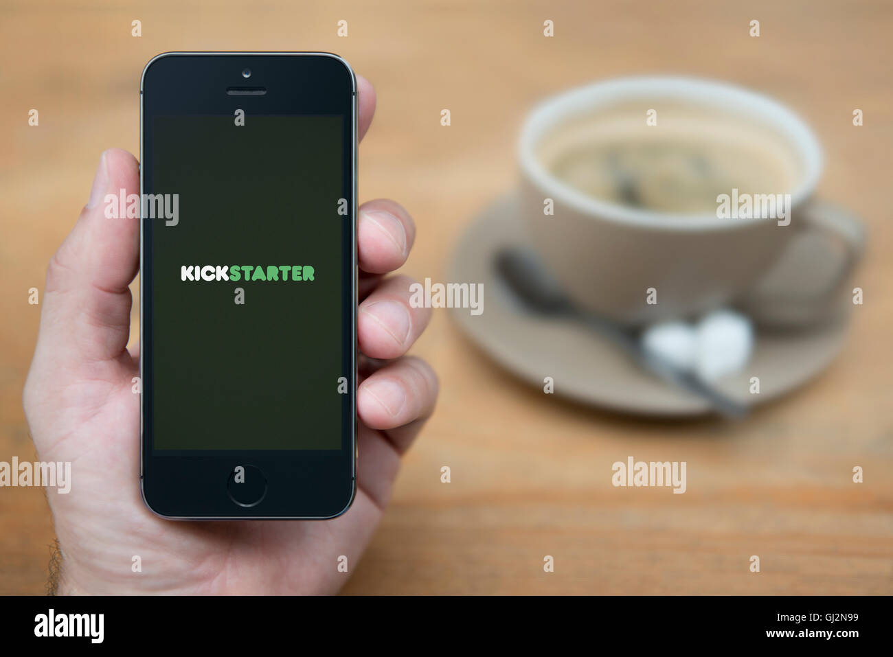 Un uomo guarda al suo iPhone che visualizza il logo Kickstarter, mentre sat con una tazza di caffè (solo uso editoriale). Foto Stock