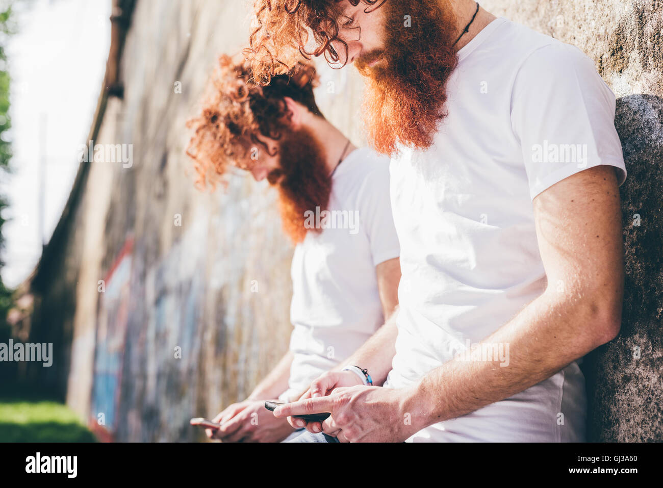 Giovane maschio hipster gemelli con la barba rossa appoggiata contro la parete texting sugli smartphone Foto Stock