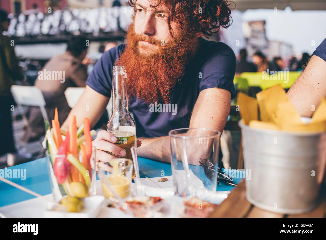 Giovane maschio hipster con rosso di capelli e la barba di bere la birra in bottiglia al marciapiede bar Foto Stock