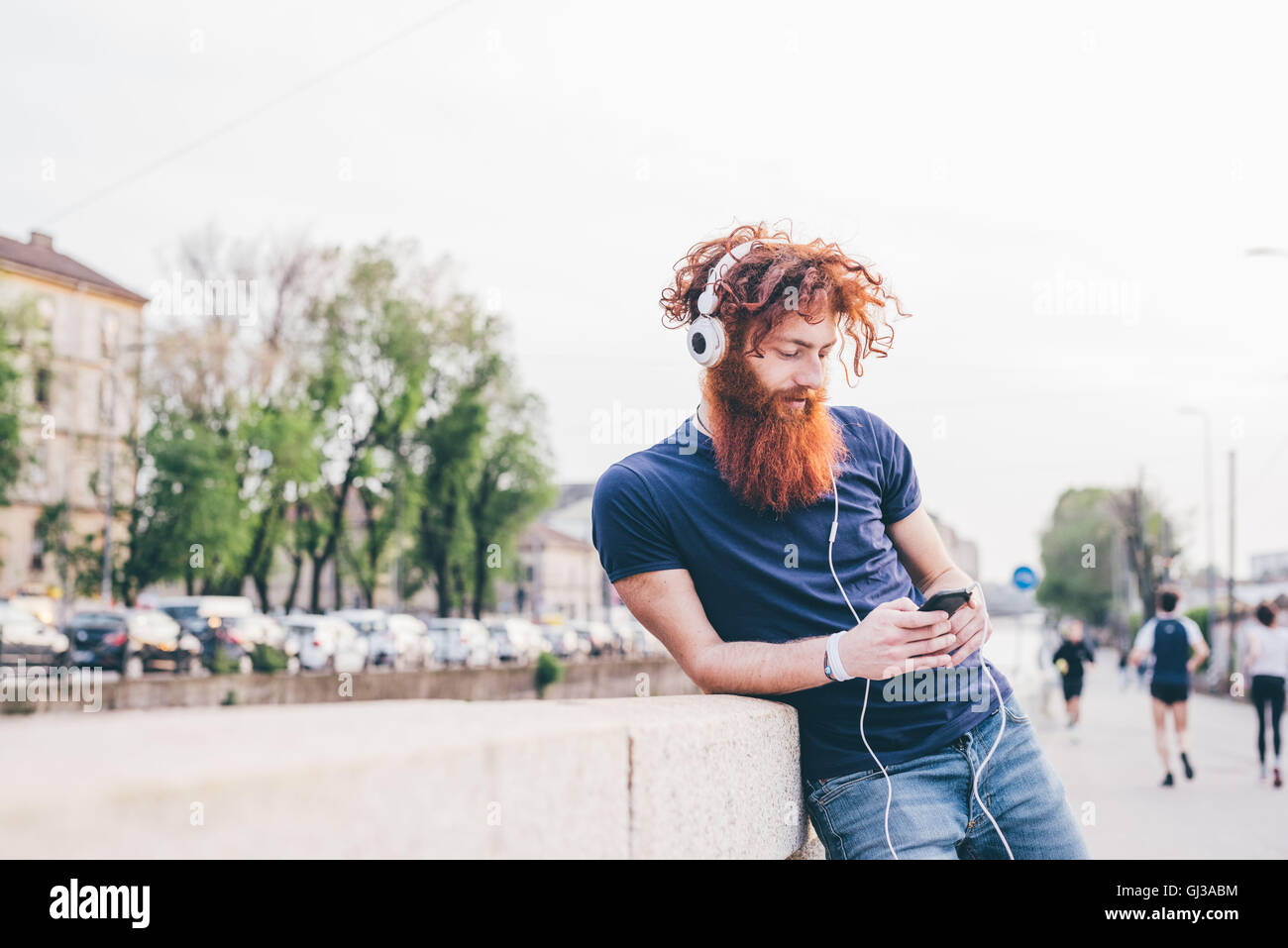 Giovane maschio hipster con capelli rossi e barba scegliendo la musica delle cuffie in città Foto Stock