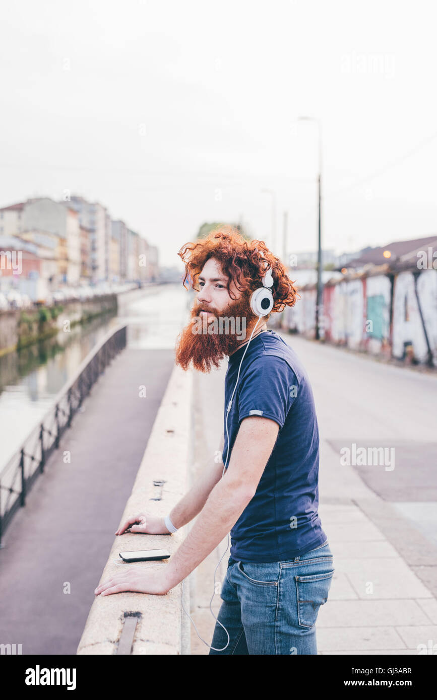 Giovane maschio hipster con i capelli rossi e la barba ascoltando le cuffie sul ponte della città Foto Stock