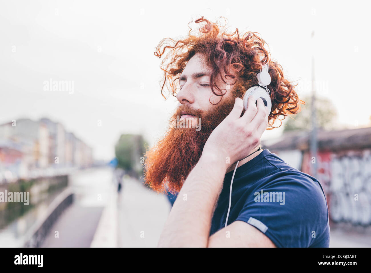 Giovane maschio hipster con i capelli rossi e la barba ascoltando le cuffie con gli occhi chiusi in città Foto Stock
