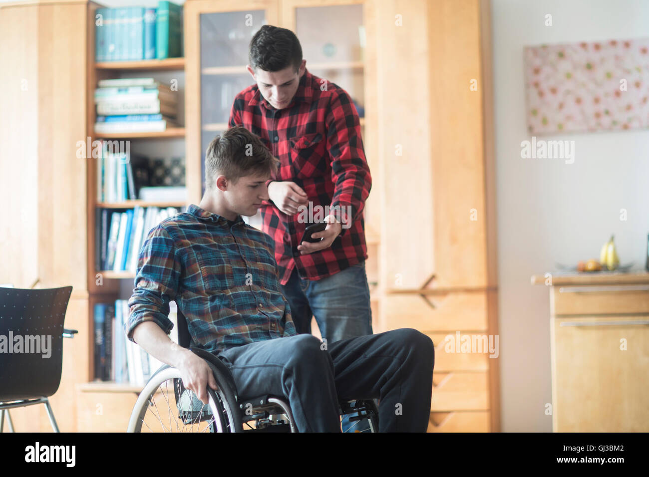 Giovane uomo con sedia a rotelle la lettura di testi di smartphone con un amico in cucina Foto Stock