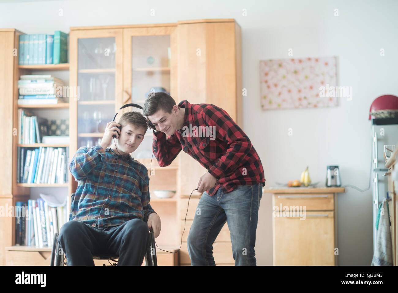 Giovane uomo con sedia a rotelle ascoltando la musica delle cuffie con un amico in cucina Foto Stock