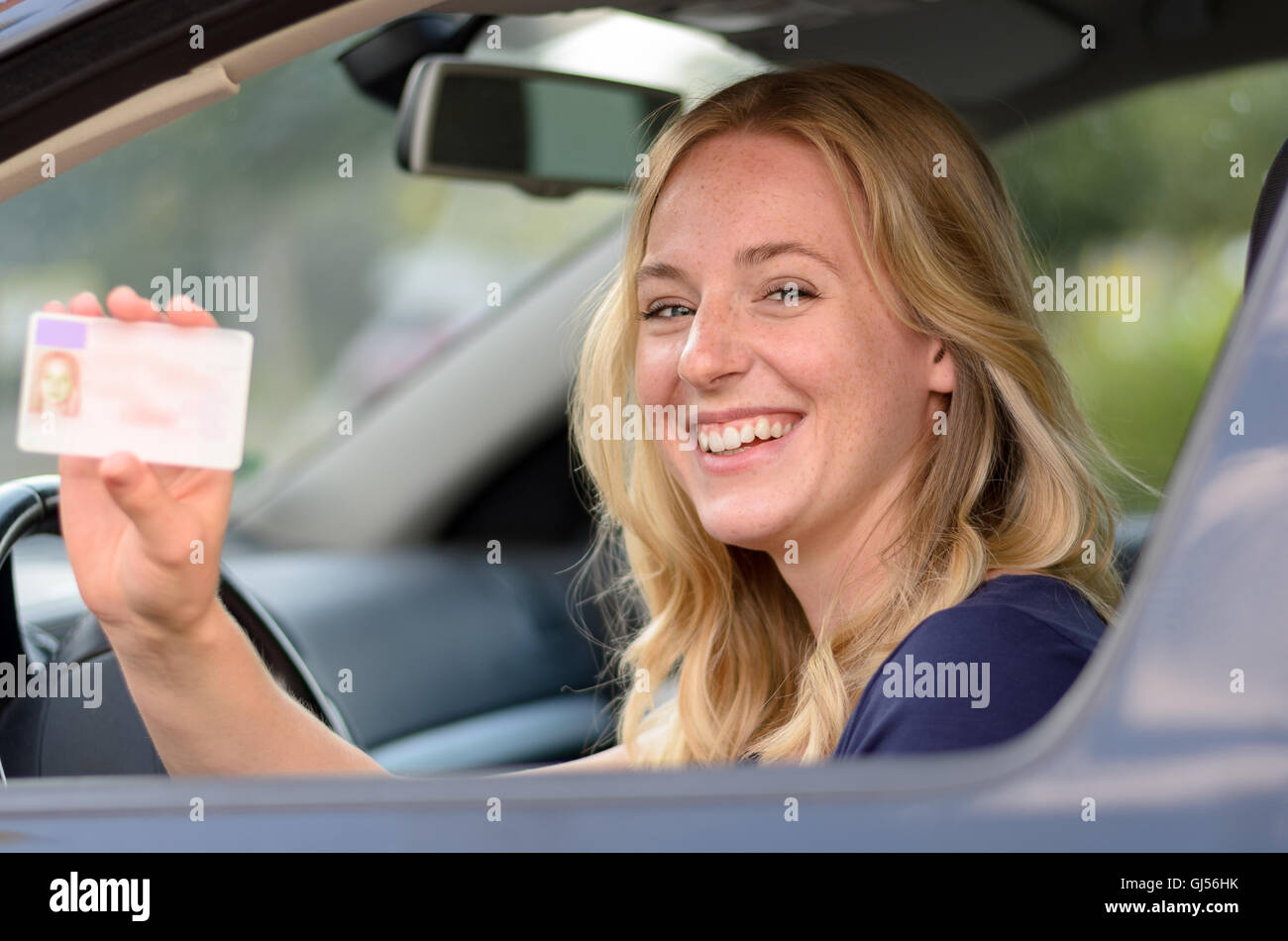 Felice giovane donna bionda seduta dietro al volante di una vettura di mostrare la sua patente di guida attraverso il finestrino aperto Foto Stock