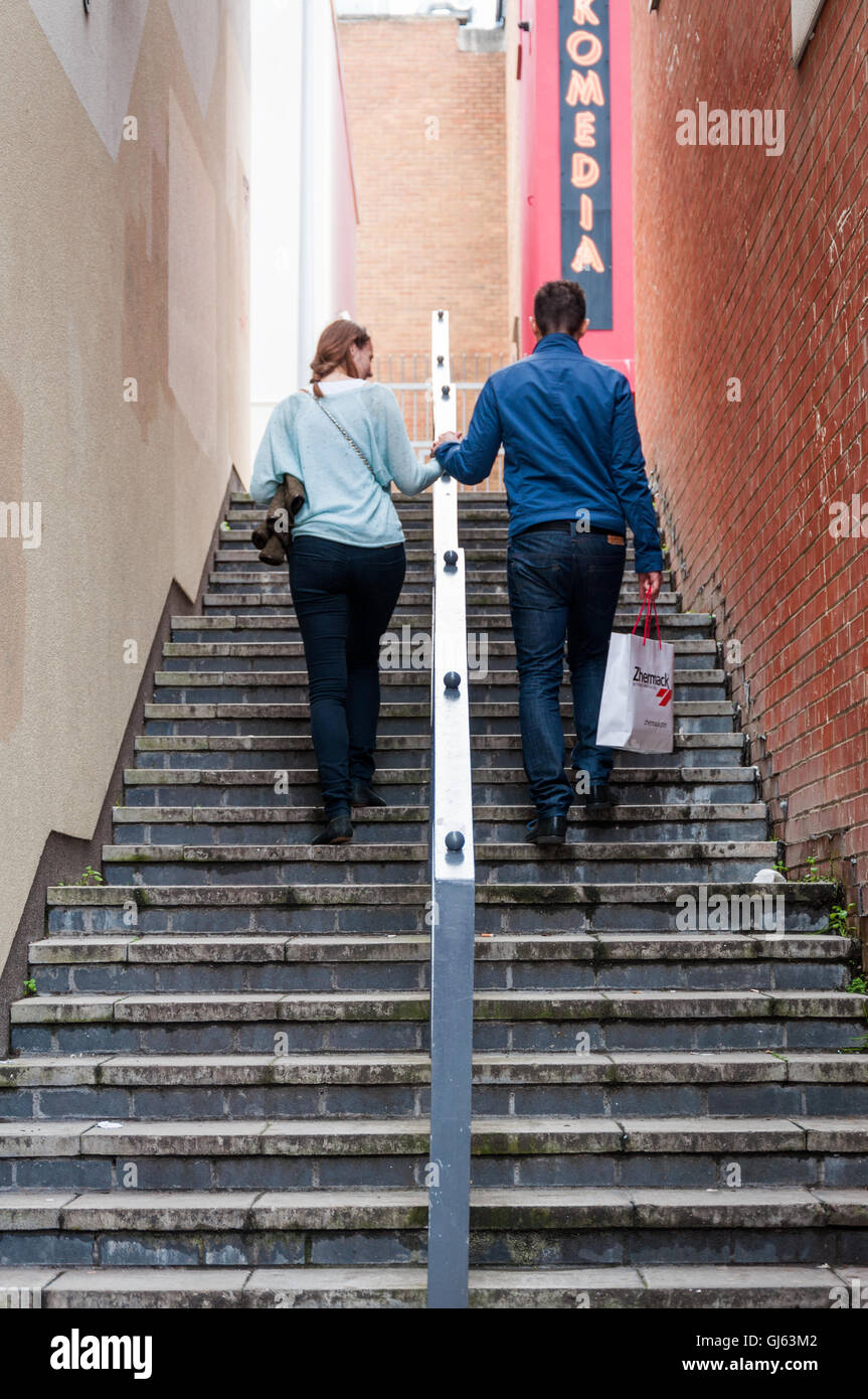 Felice coppia tenendo le mani mentre si cammina su ripide scale con il segno Komedia sullo sfondo come parodia o commedia nella vita. Brighton, Regno Unito Foto Stock