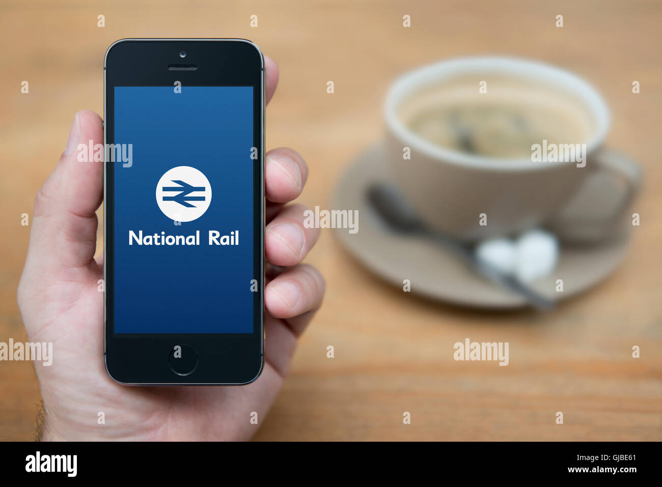 Un uomo guarda al suo iPhone che visualizza il National Rail logo, mentre sat con una tazza di caffè (solo uso editoriale). Foto Stock
