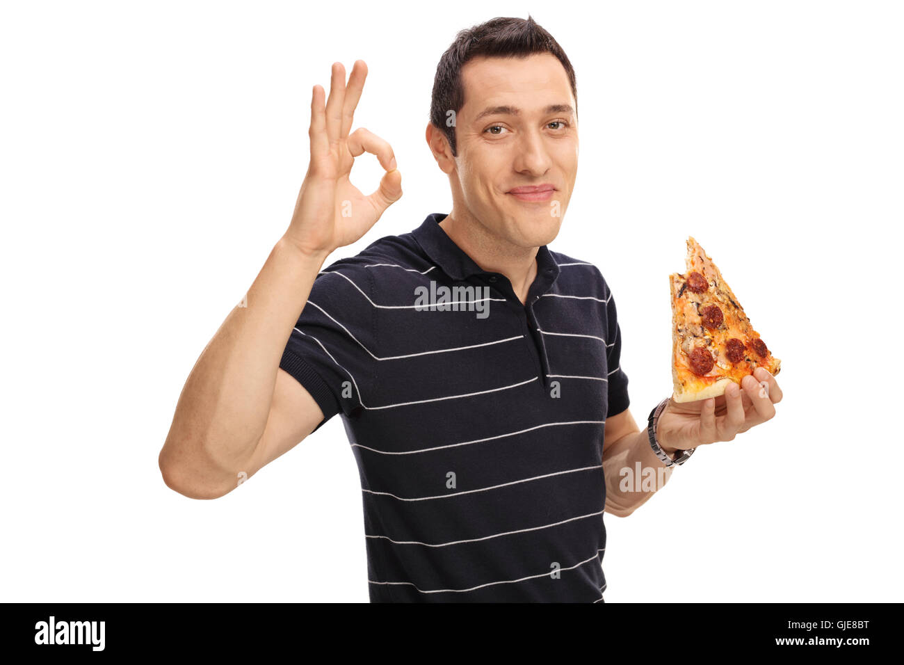 Gioiosa giovane mangiare una fetta di pizza e facendo un gesto ok con la sua mano isolati su sfondo bianco Foto Stock