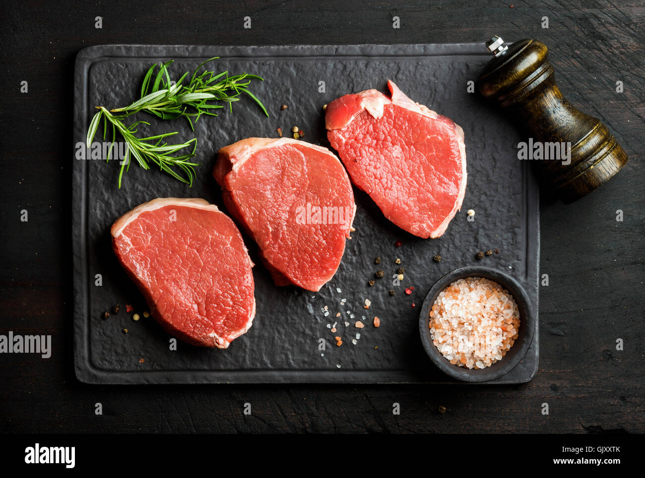 Carne di manzo crudo occhio Round bistecche con spezie e rosmarino su ardesia nera pietra oltre il bordo di legno scuro, sfondo vista superiore, orizzontale Foto Stock