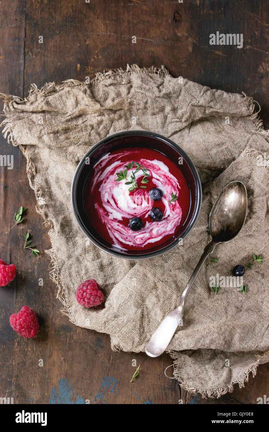 Nero vaso di ceramica di lampone dolce zuppa con crema, servito con il cucchiaio, frutti di bosco freschi e timo su un letto di sacco su legno vecchio t Foto Stock