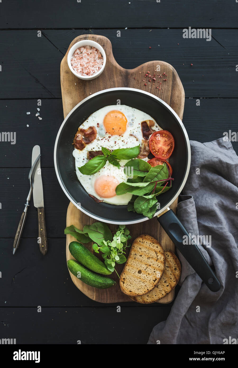 Pan di uova fritte, pancetta, pomodori ciliegia e erbe aromatiche fresche con il pane sulla tavola di legno scuro su sfondo di legno, vista dall'alto Foto Stock