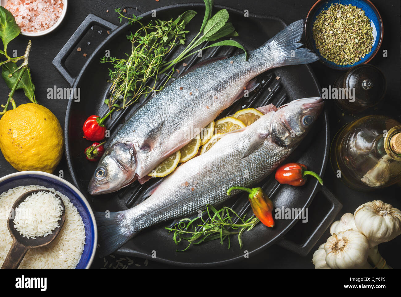 Ingredienti per cookig pesci sani la cena. Materie non cotti spigola pesce con riso, limone, erbe e spezie su nero ferro grill Foto Stock