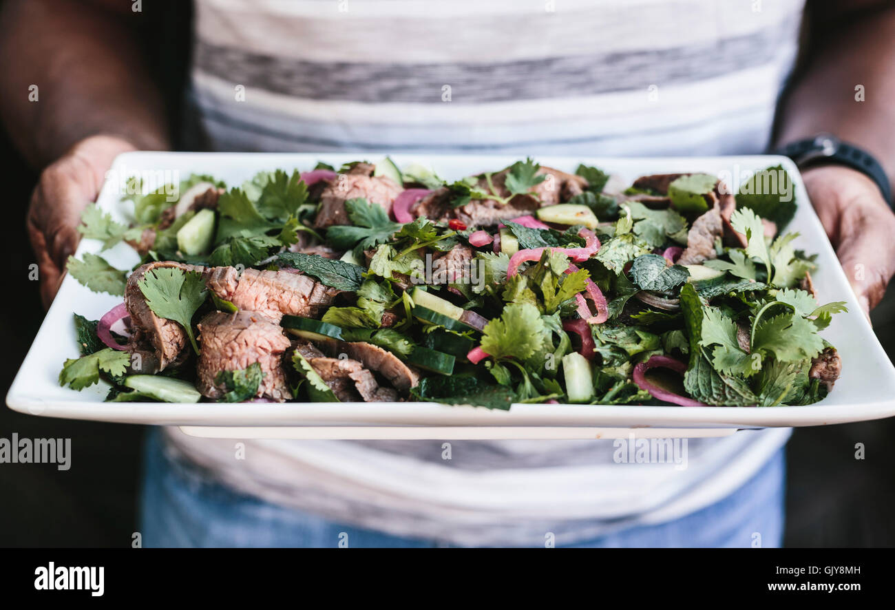Un uomo è in possesso di una piastra di Thai manzo alla griglia e insalata di erbe. Foto Stock