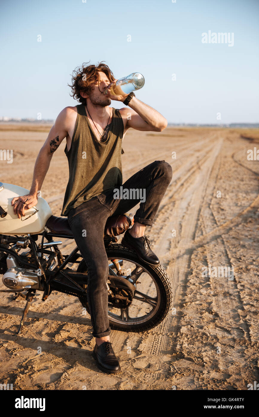 Giovane uomo brutale seduta sulla sua moto nel deserto e acqua potabile Foto Stock