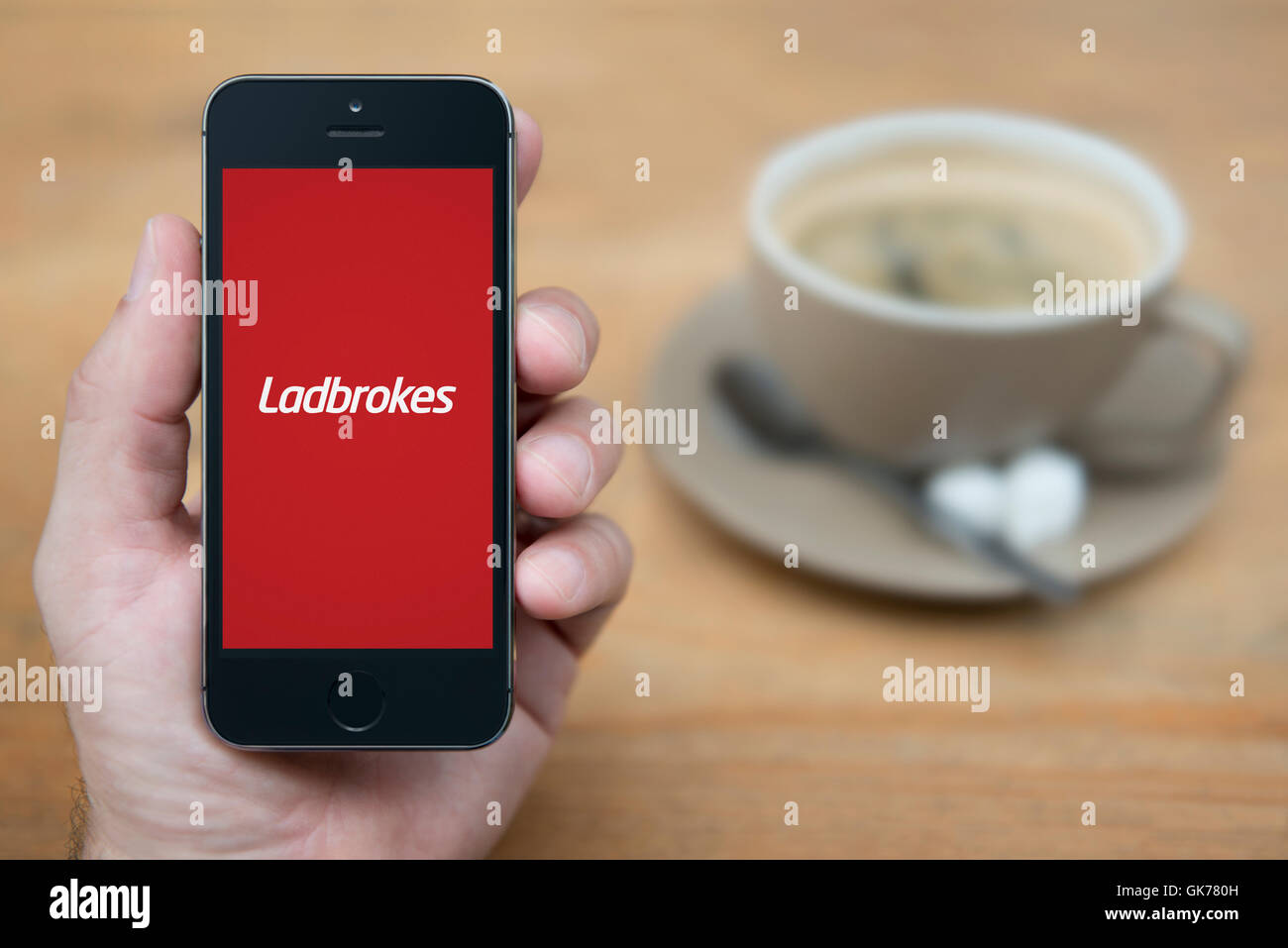 Un uomo guarda al suo iPhone che visualizza il logo Ladbrokes, mentre sat con una tazza di caffè (solo uso editoriale). Foto Stock