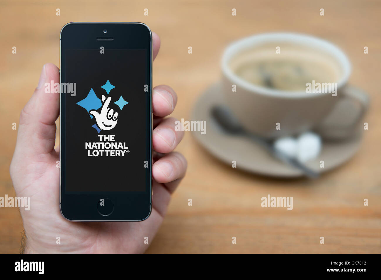 Un uomo guarda al suo iPhone che visualizza la lotteria nazionale logo, mentre sat con una tazza di caffè (solo uso editoriale). Foto Stock
