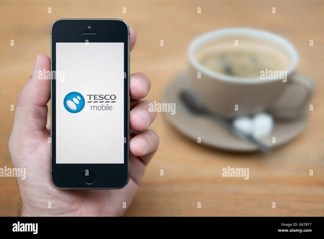 Un uomo guarda al suo iPhone che visualizza il Tesco Mobile logo, mentre sat con una tazza di caffè (solo uso editoriale). Foto Stock
