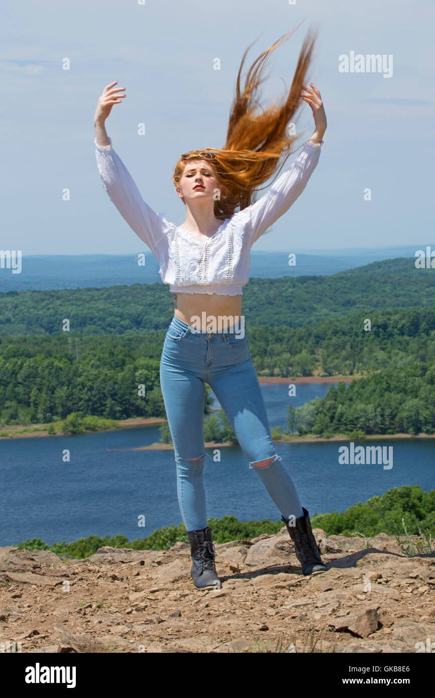 Bella testa rossa in skinny jeans, stivali neri e top bianco, sulla cima della montagna con il vento nei capelli e un lago sottostante. Foto Stock