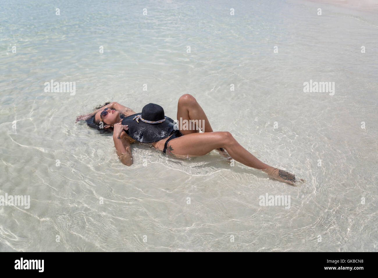 La donna in nero di un costume da bagno con spiaggia nera hat posa nelle acque di Osprey Cay, Exuma Cays, Isole Bahamas Foto Stock