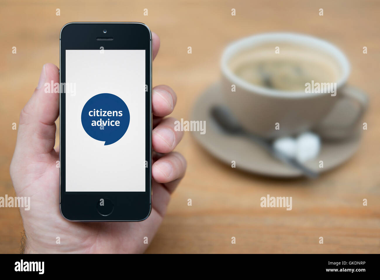 Un uomo guarda al suo iPhone che visualizza il Citizens Advice logo, mentre sat con una tazza di caffè (solo uso editoriale). Foto Stock