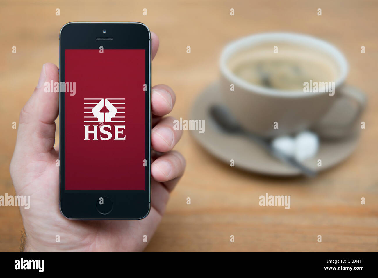 Un uomo guarda al suo iPhone che visualizza il logo HSE, mentre sat con una tazza di caffè (solo uso editoriale). Foto Stock