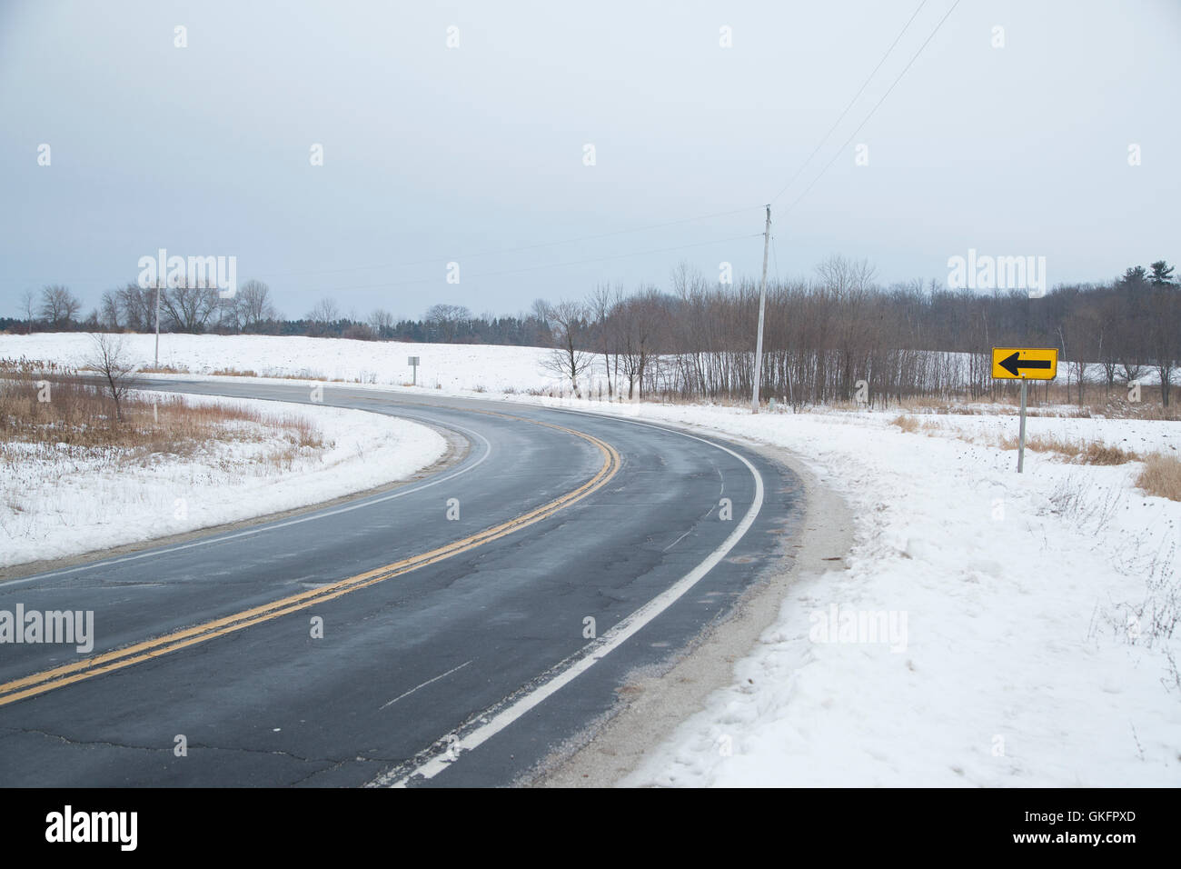 A due corsie asfalto road accanto a una coperta di neve delle curve di campo passato un segno di freccia sul grigio di una giornata invernale Foto Stock