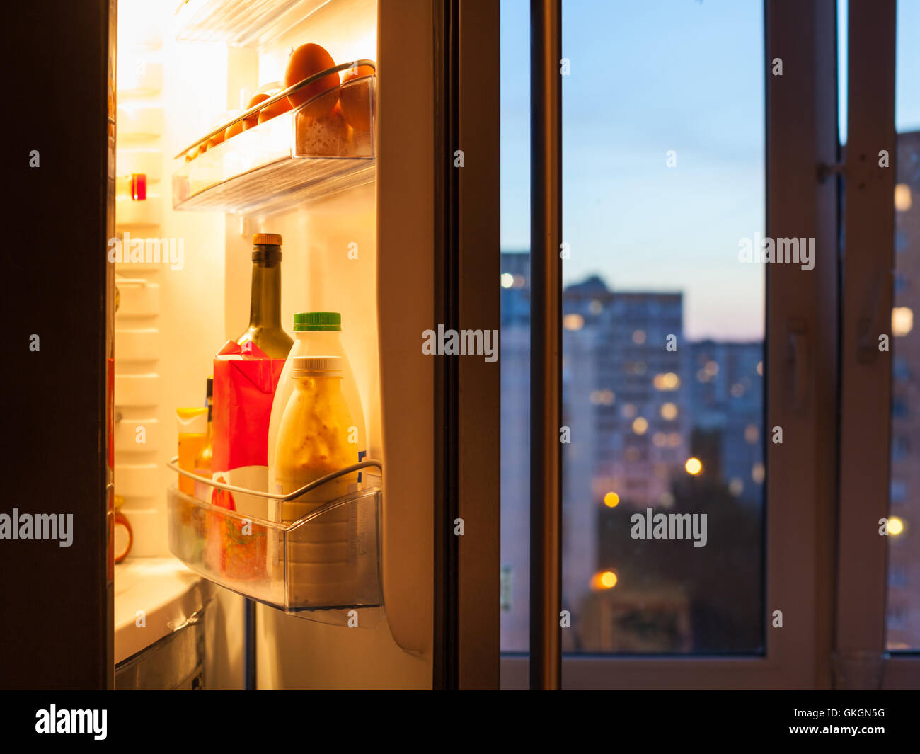 Aprire lo sportello del frigorifero con il cibo e la vista della città attraverso la finestra di casa in serata Foto Stock