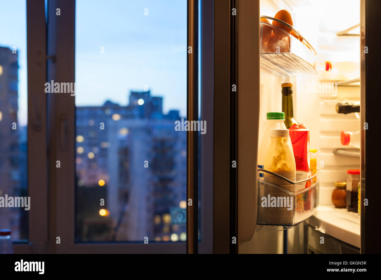 Aprire lo sportello del frigorifero con pasto e vista della città attraverso la finestra in sera Foto Stock