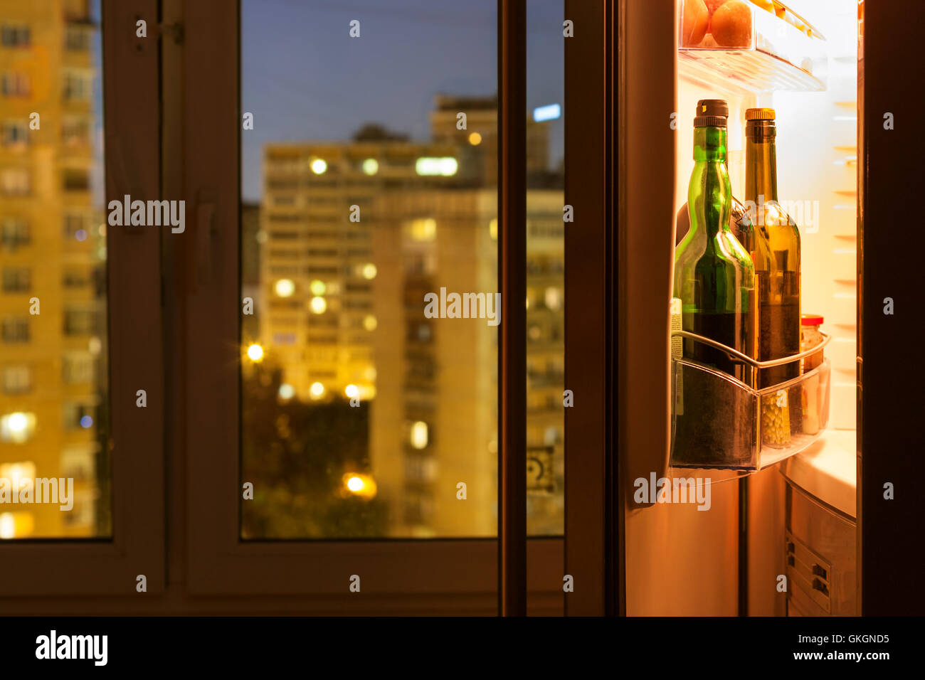 Aprire lo sportello del frigorifero con bottiglie di vino e la vista della città attraverso la finestra di casa a notte Foto Stock
