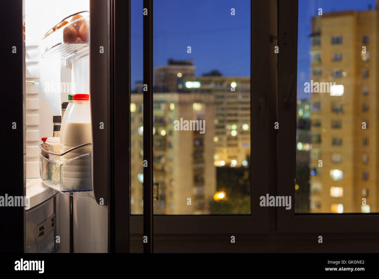 Aprire lo sportello del frigorifero con bottiglie di latte e la vista della città attraverso il vetro di una finestra nella notte Foto Stock