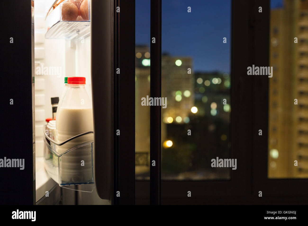Aprire lo sportello del frigorifero domestico con bottiglie di latte e la vista della città attraverso la finestra nella notte Foto Stock