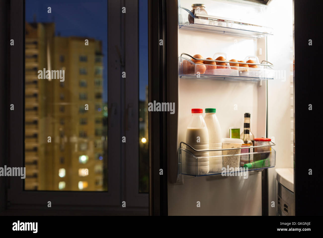 Aprire lo sportello del frigorifero domestico con prodotti caseari e vista della città attraverso la finestra nella notte Foto Stock