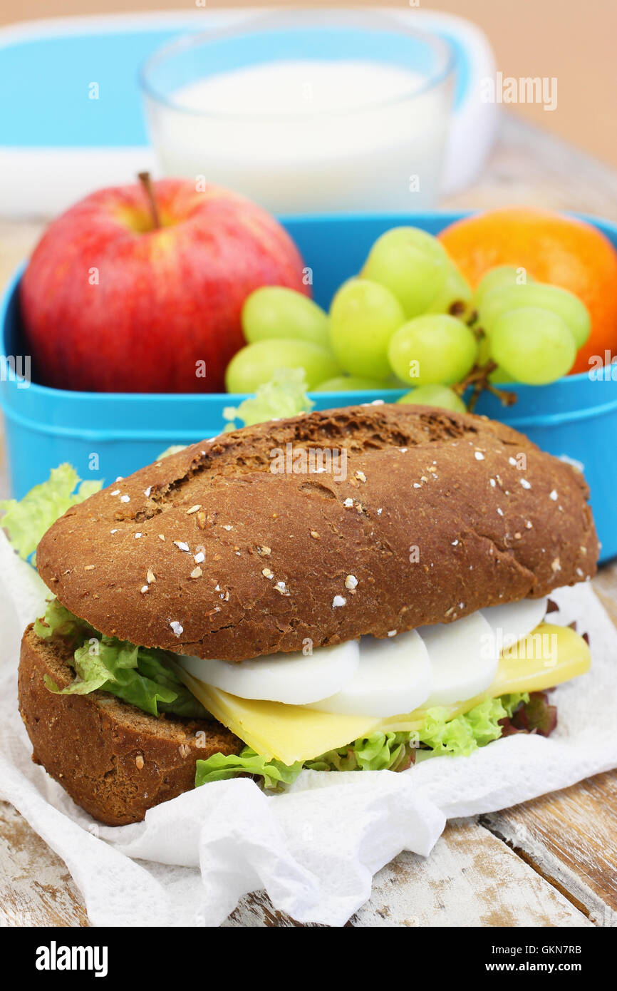Marrone sandwich di pane con formaggio, uovo sodo e lattuga, scatola di pranzo pieno di frutta fresca e un bicchiere di latte Foto Stock