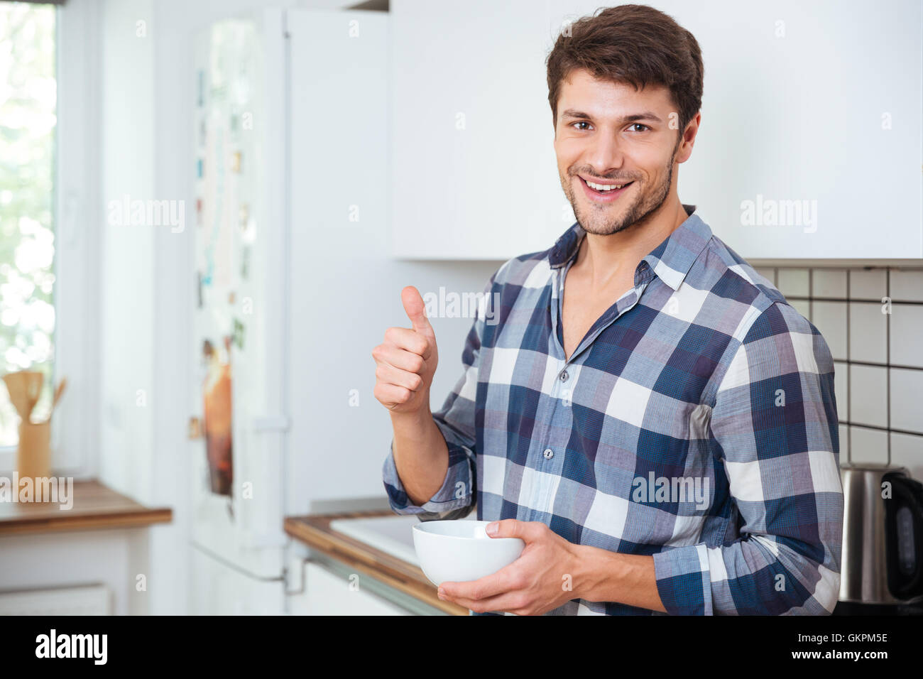 Allegro Giovane Uomo in camicia a scacchi permanente sulla cucina e mostrando pollice in alto Foto Stock