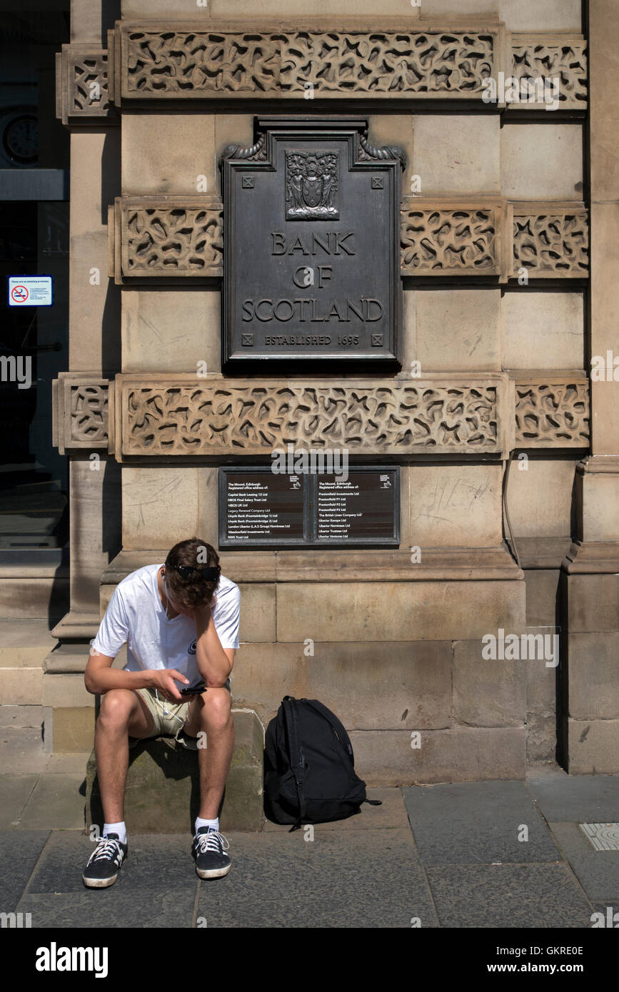 Un giovane uomo controlla il suo telefono mentre è seduto alla porta della Lloyds Banking Group Sede scozzese ex HBoS Foto Stock