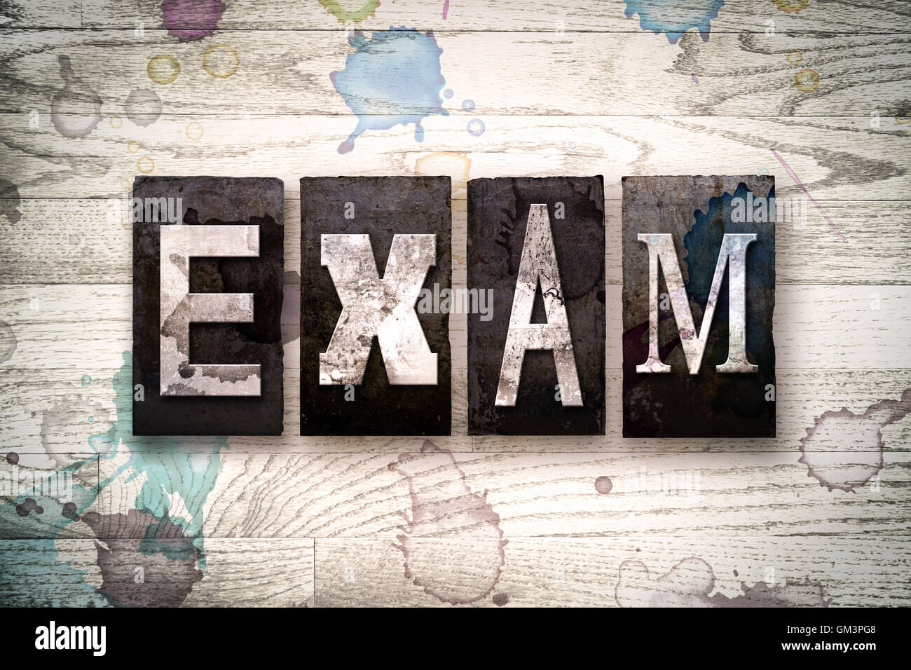 The word "EXAM" written in vintage dirty metal letterpress type on a whitewashed wooden background with ink and paint stains. Foto Stock