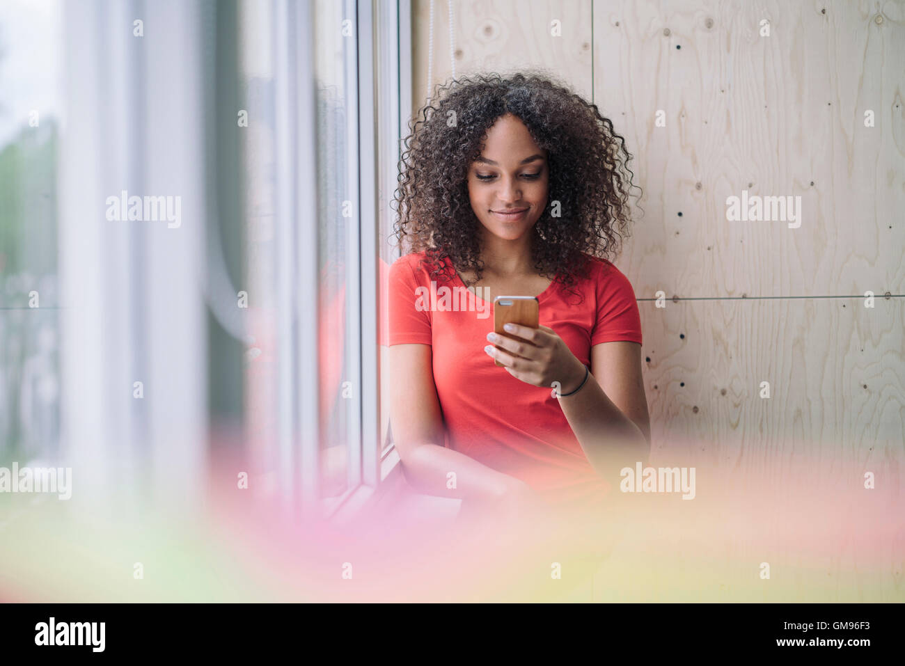 Giovane donna in piedi dalla finestra, utilizzando smart phone Foto Stock