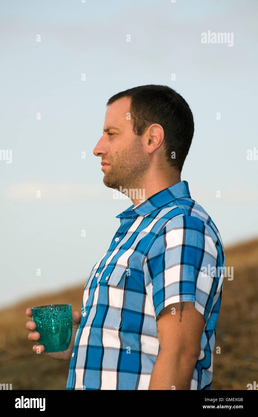 Uomo in piedi e guardare fuori sul pendio di una collina tenendo un bicchiere in mano che indossa un plaid shirt. Foto Stock