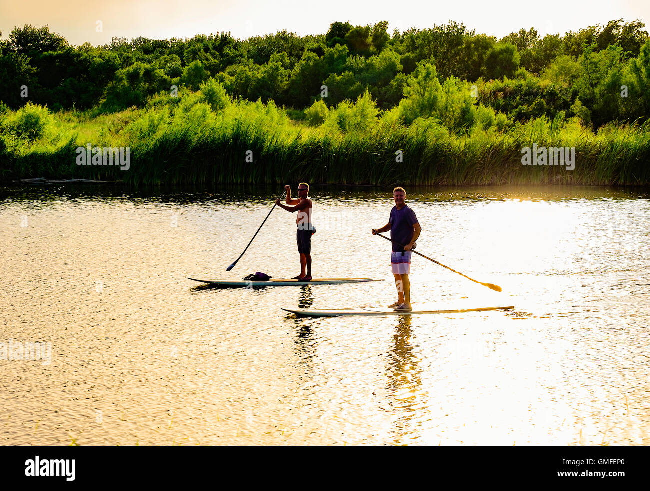 Due Caucasian uomini di mezza età oar stand up paddleboards giù il nord del fiume canadese. Tramonto riflesso, Oklahoma, Stati Uniti d'America. Foto Stock