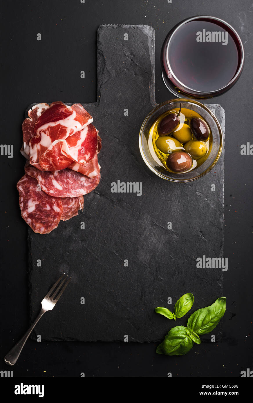 Bicchiere di vino rosso, carne antipasto, olive e basilico sul nero ardesia scheda di pietra su sfondo scuro Foto Stock