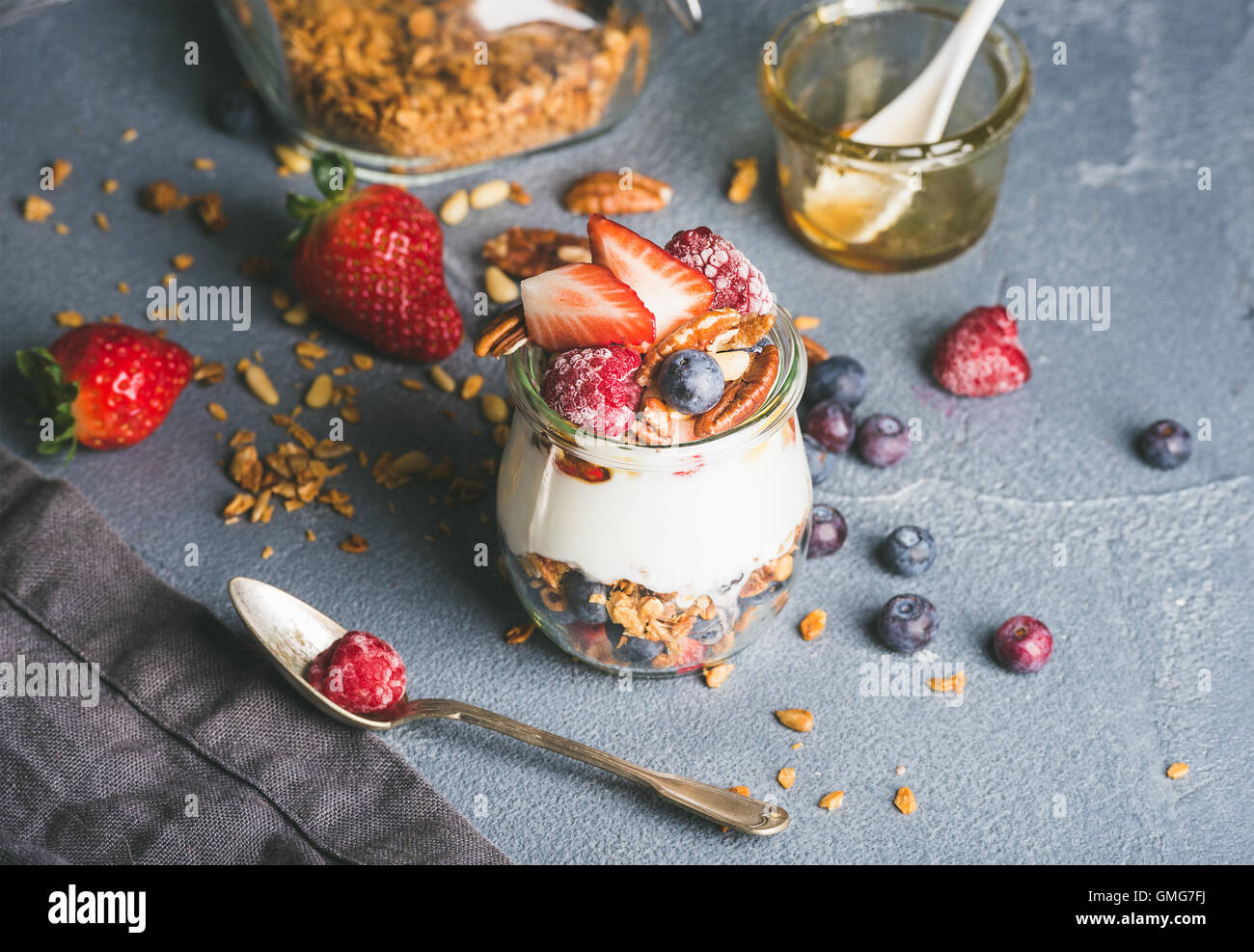 Yogurt oat granola con frutti di bosco freschi, noci, miele e foglie di menta in un barattolo di vetro sul cemento grigio Sfondo testurizzata Foto Stock