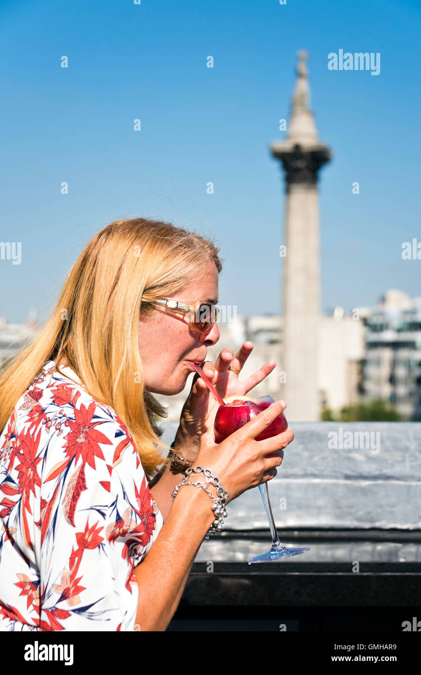 Ritratto verticale di una donna gustando un cocktail affacciato su Nelson's colonna in Trafalgar Square, Londra, sotto il sole. Foto Stock